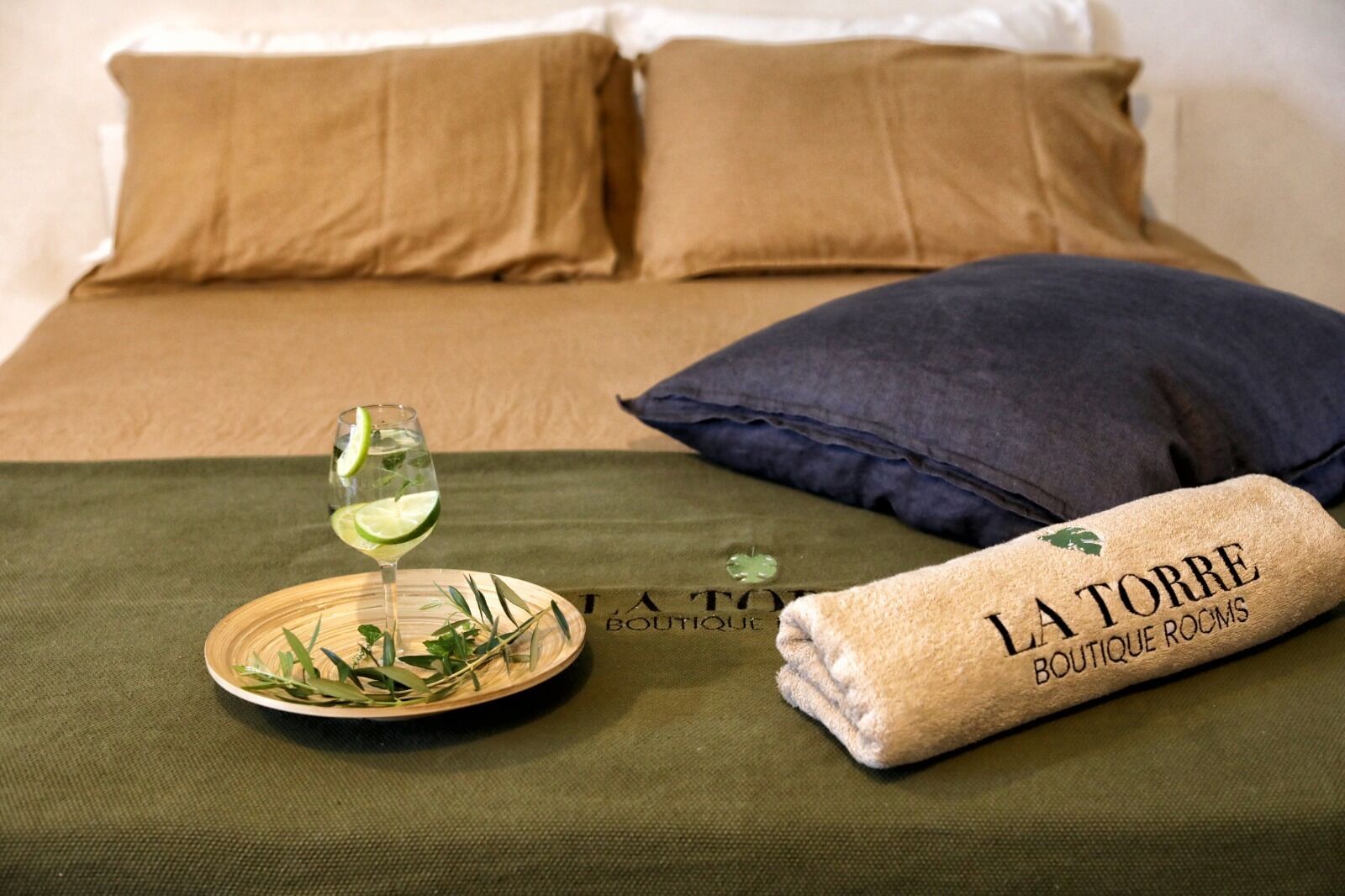 A neatly made bed with a green bedspread, a folded towel with "LA TORRE BOUTIQUE ROOMS" text, and a glass of white wine with lime garnish on a wooden tray.