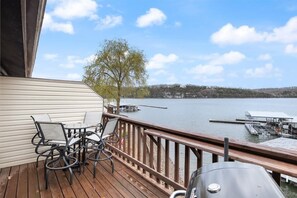 Um deck com mobiliário externo e churrasqueira, com vista para um lago com docas e uma árvore.