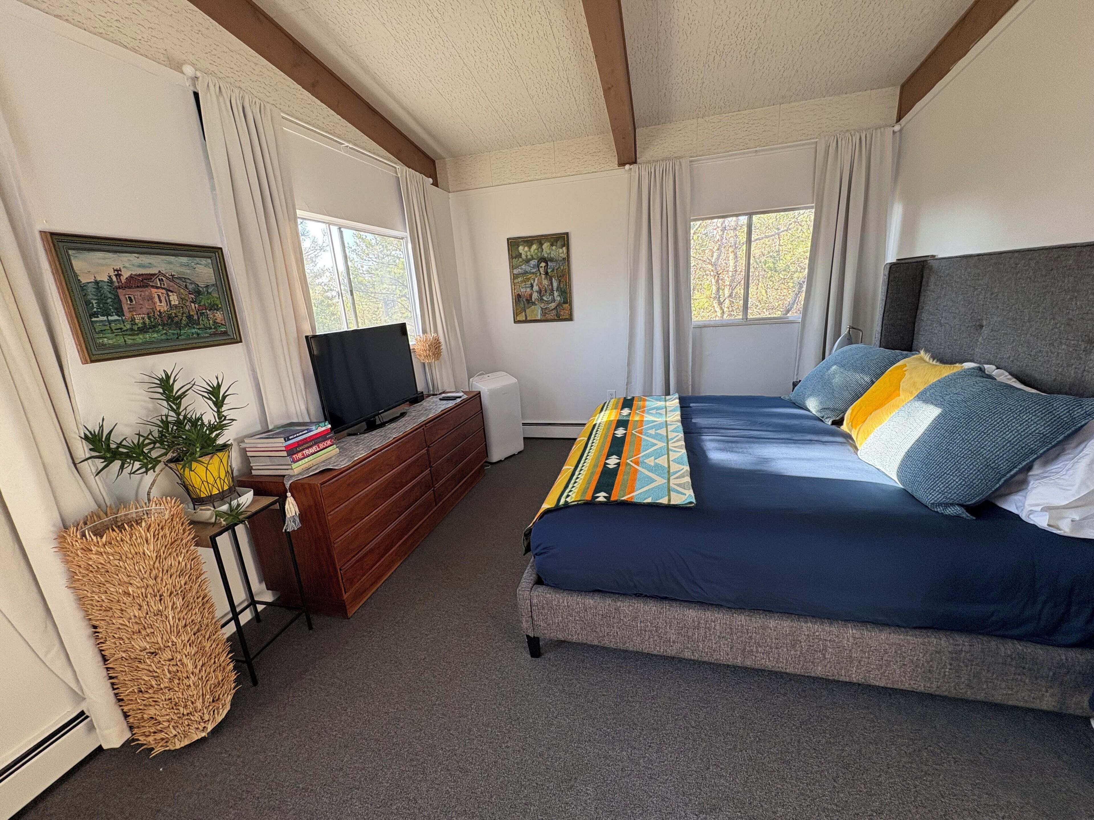 Quarto com cama, penteadeira, televisão e duas janelas com cortinas.