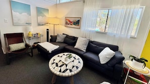 A modern living room with a sectional sofa, a round coffee table, a side chair, and a floor lamp. There are two windows with sheer curtains, a painting on the wall, and a bookshelf with books and decorative items.