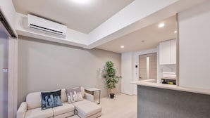 A modern living room with a beige sectional sofa, a small side table, a potted plant, and a built-in kitchen counter. - MP Hotel Hakata (Fukuoka)