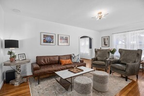 Uma sala de estar com um sofá de couro marrom, uma mesa de centro, duas poltronas, uma pequena mesa lateral com abajur e quadros nas paredes.