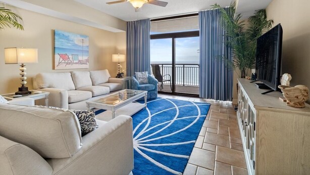A spacious living room with a ceiling fan, a large window with curtains, a flat-screen TV, and a blue patterned rug. - Sun, Sand and Sea Views 3 Bdrm Condo with Balcony (North Myrtle Beach)