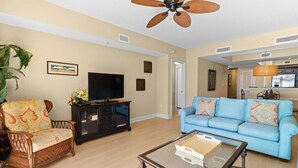 A living room with a television, a blue sofa, a wicker chair, and a glass coffee table.