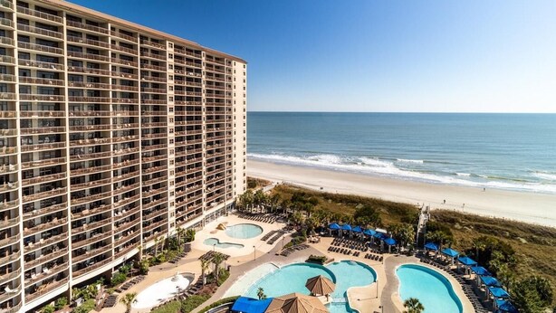 Ein Hochhotel mit mehreren Balkonen mit Blick auf einen Strand und einen Poolbereich mit Liegestühlen und Sonnenschirmen.