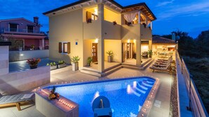 Una casa de dos pisos con piscina, mobiliario exterior y una entrada bien iluminada.
