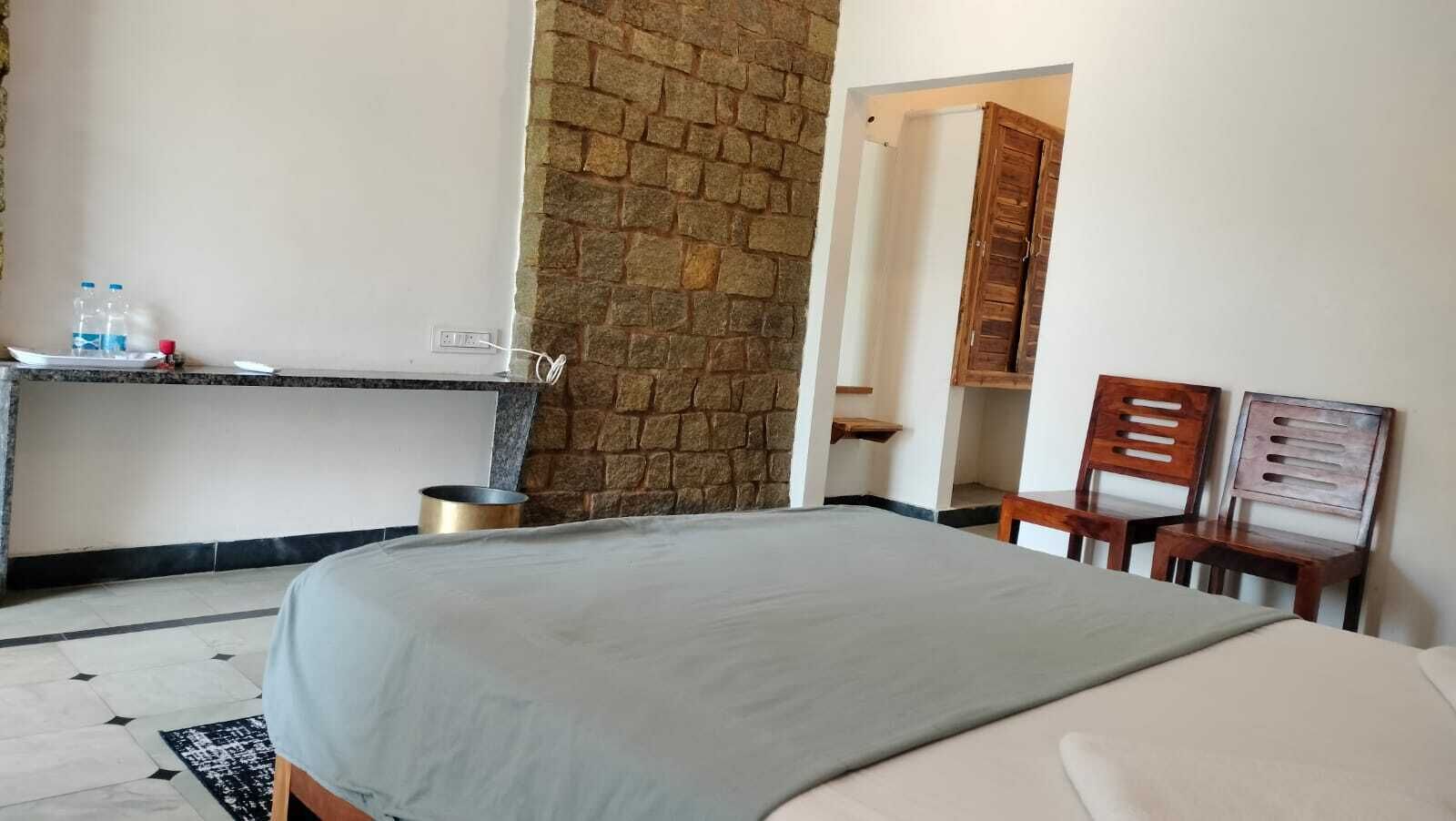 Habitación de hotel con cama, una mesita con un recipiente metálico, pared de ladrillo y dos sillas de madera.
