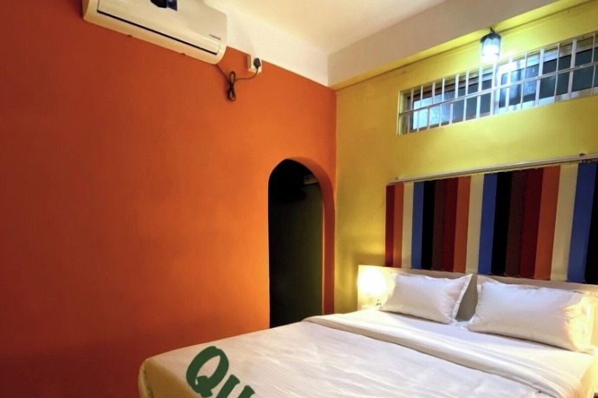 A hotel room with a bed labeled "Queen Size," a window with vertical blinds, an air conditioning unit, and a TRIDHARARA logo.
