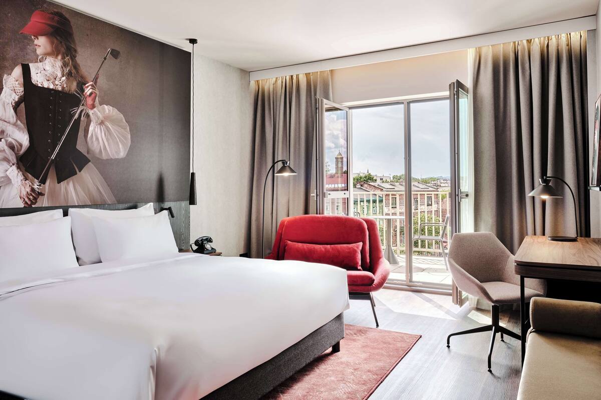 Habitación de hotel con una cama grande, una silla roja, un escritorio y vistas a edificios a través de la ventana.