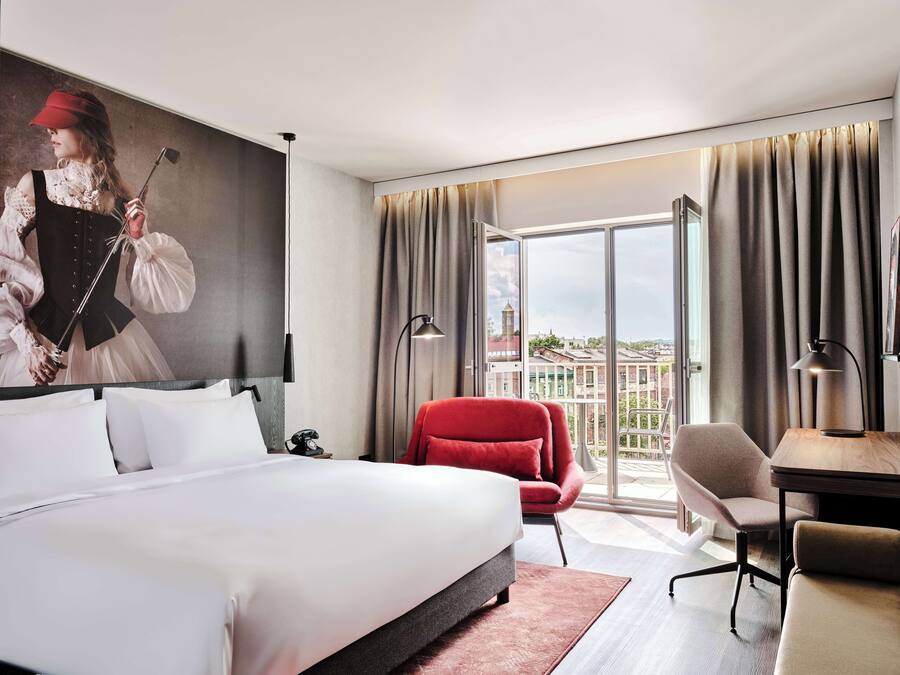 Habitación de hotel con una cama grande, una silla roja, un escritorio y vistas a edificios a través de la ventana.