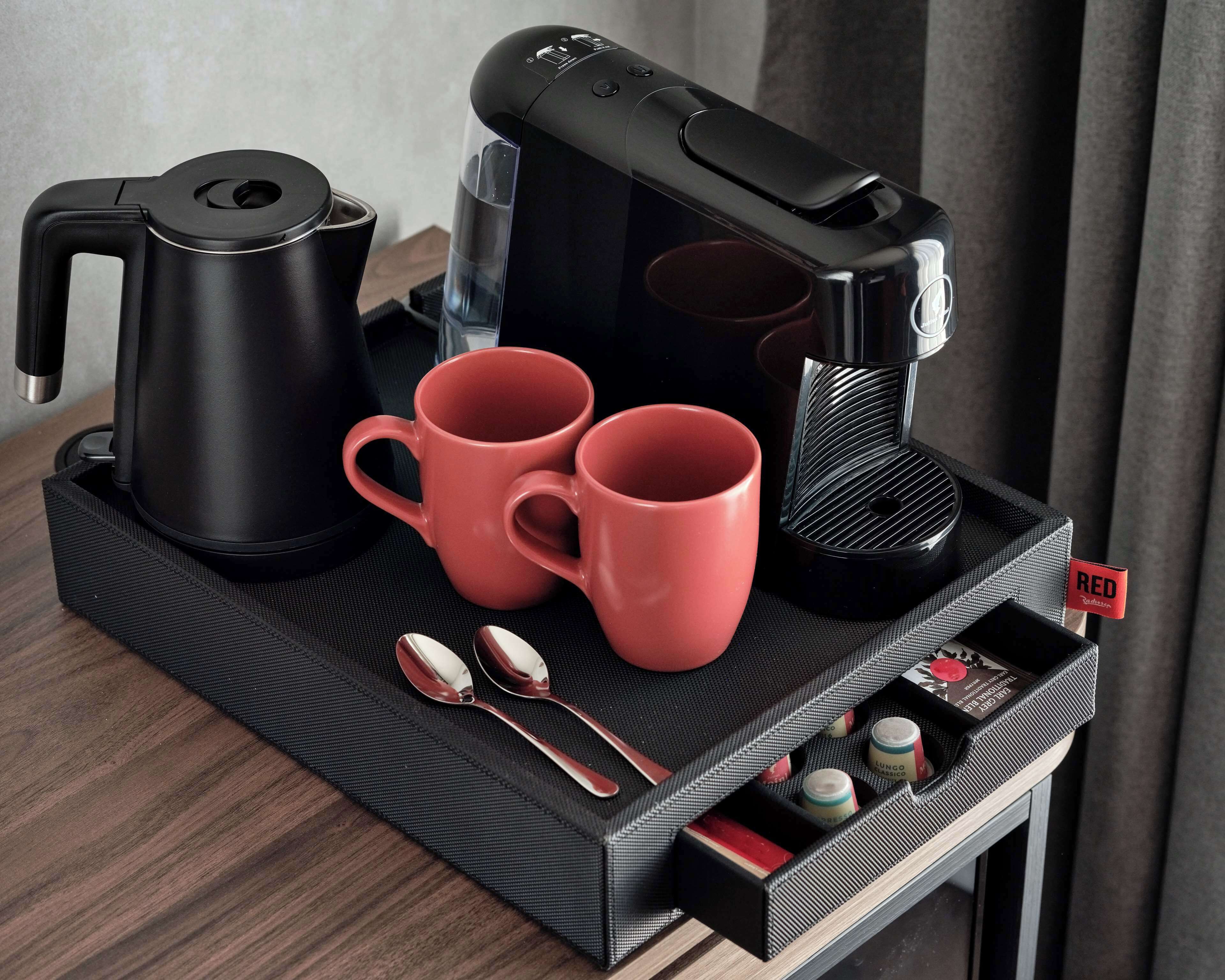 A black tray with a coffee maker, two red mugs, a black kettle, and a spoon set on a textured mat.