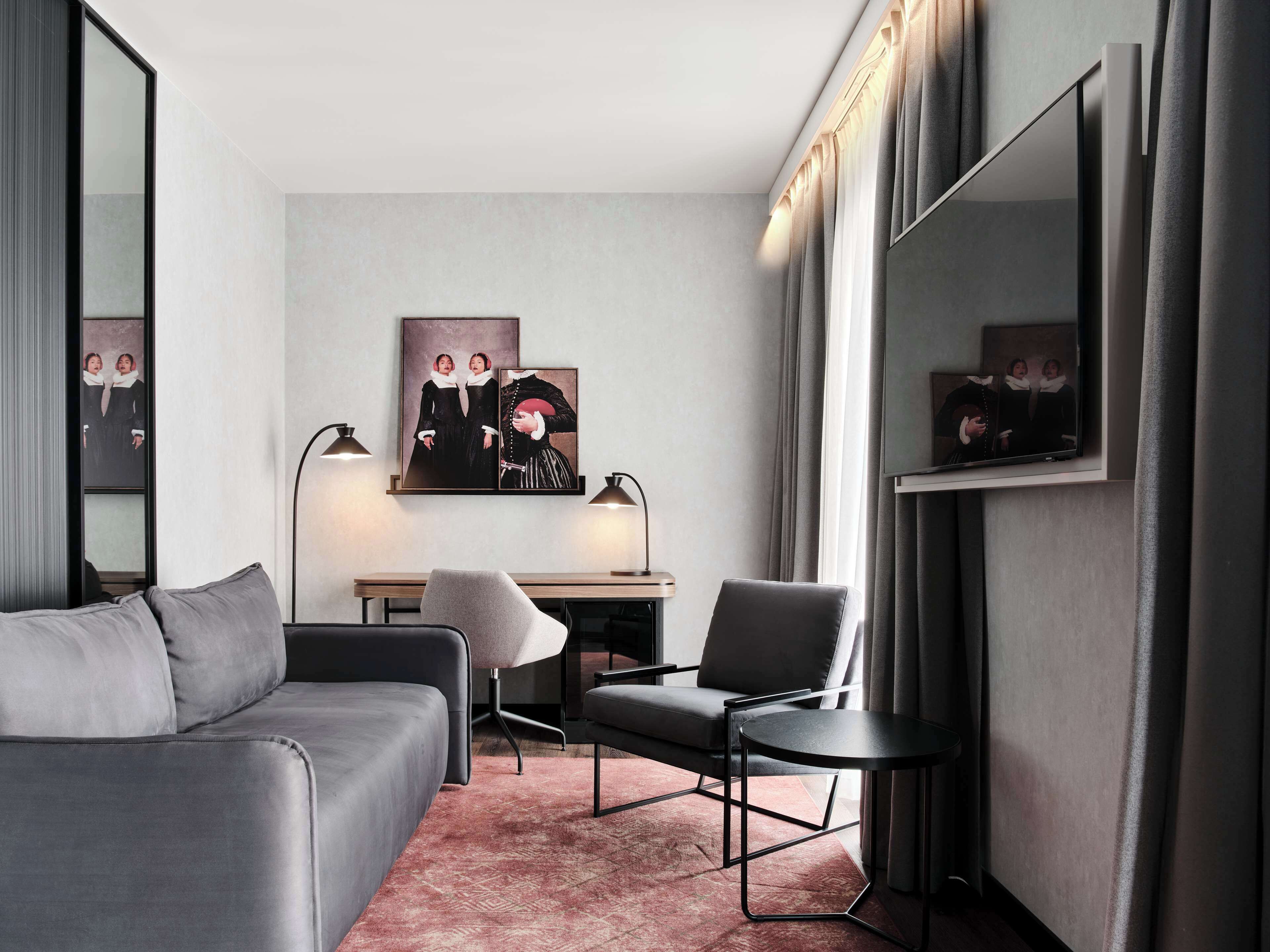 Habitació d'hotel moderna amb un sofà gris, una taula rodona petita, una escriptori amb una cadira i obra d'art a la paret.