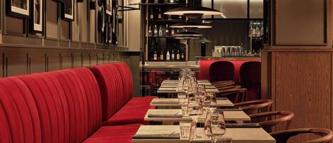 Un restaurante con cabinas tapizadas en rojo, mesas ordenadas con copas de vino y un área de bar al fondo.