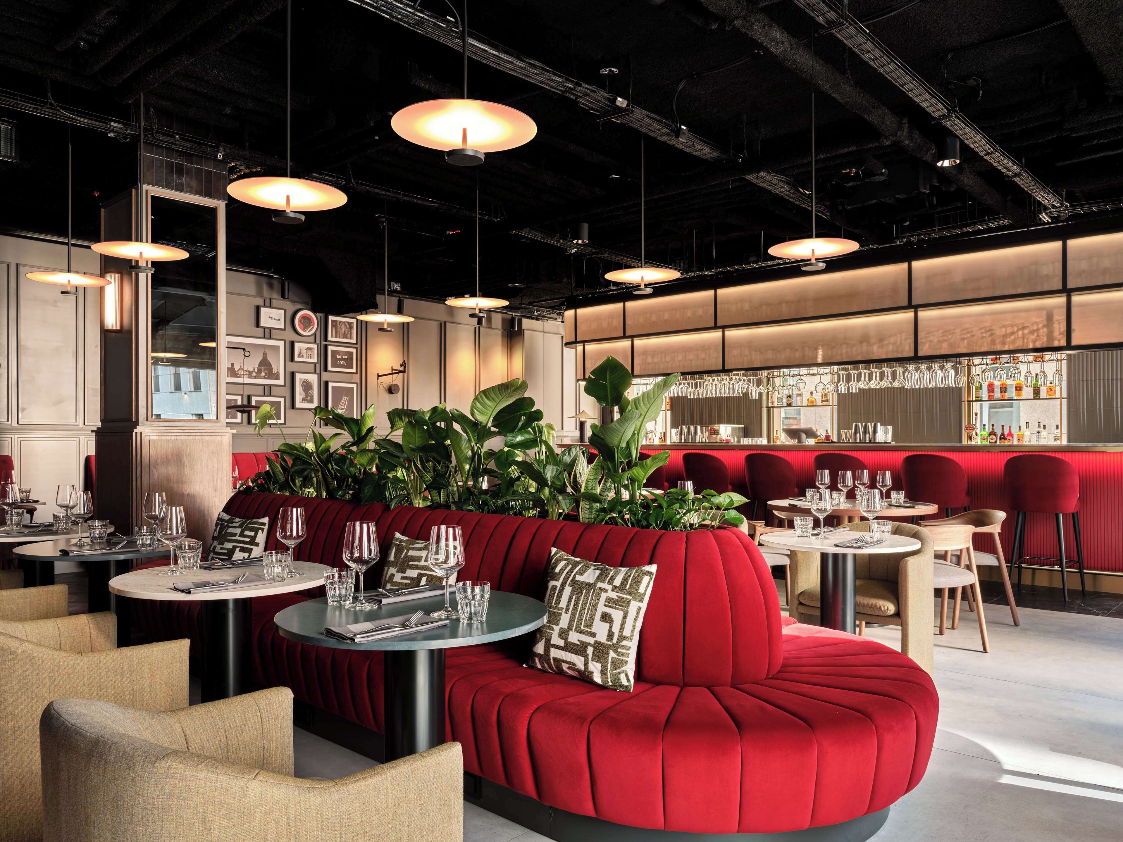 A modern restaurant interior with round tables, red upholstered seating, hanging pendant lights, and a bar area with glassware.