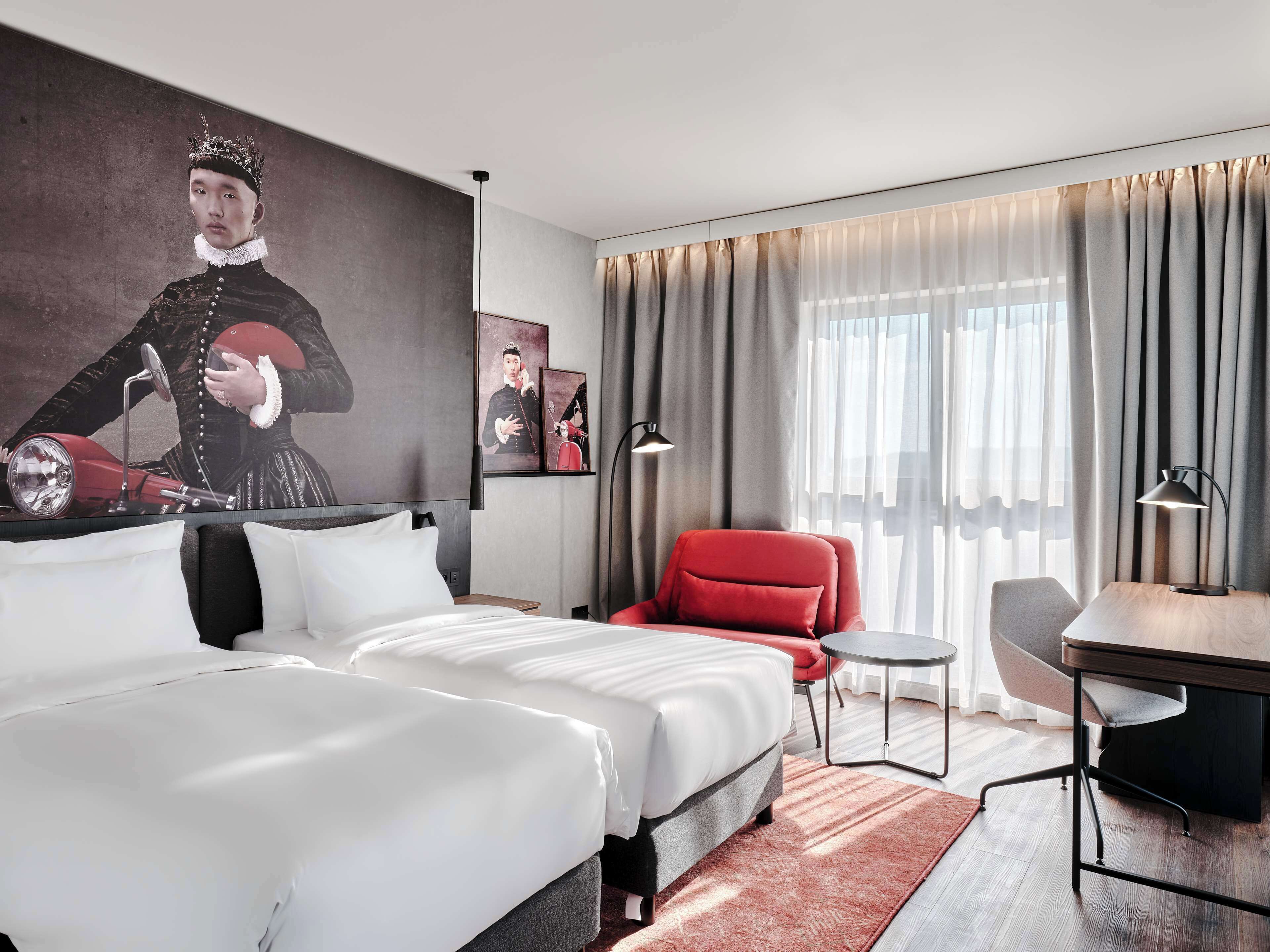 A hotel room with two beds, a red armchair, a small table, and a desk with a lamp. There are curtains and a painting on the wall.