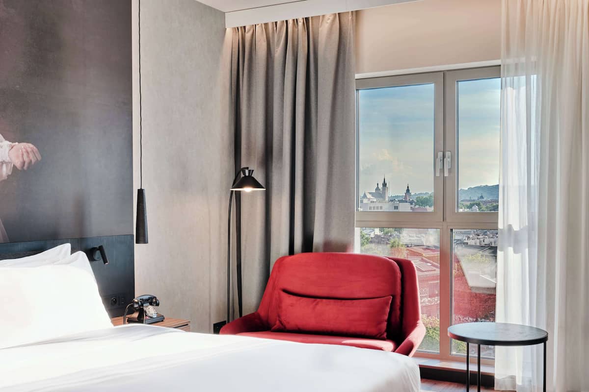 Habitación de hotel con una cama, una silla roja, una mesita, una lámpara y un ventanal con vistas a la ciudad.