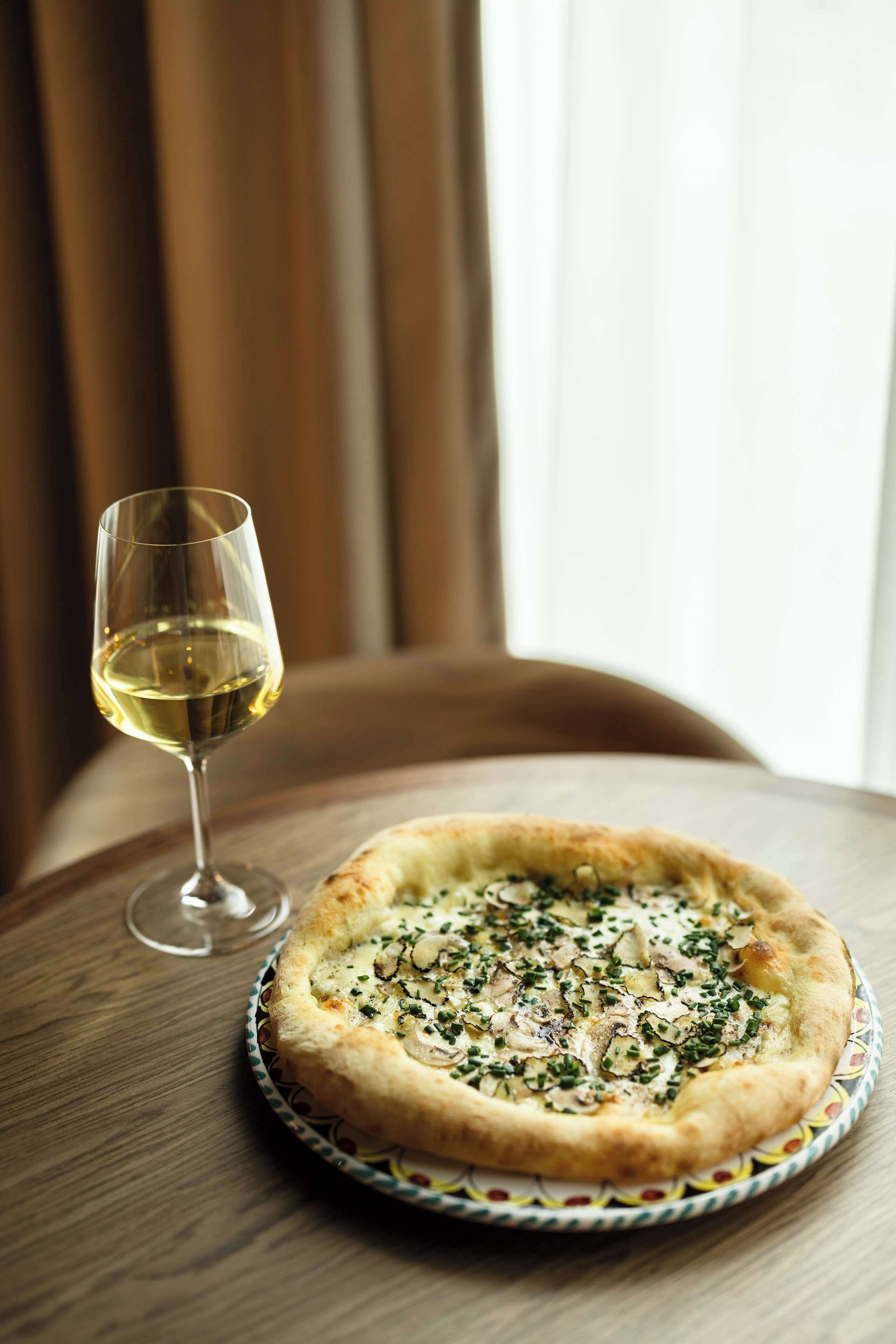 Una pizza en un plato con una copa de vino blanco sobre una mesa de madera.