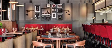A modern restaurant interior with round tables, red upholstered booths, and a wall adorned with framed pictures.