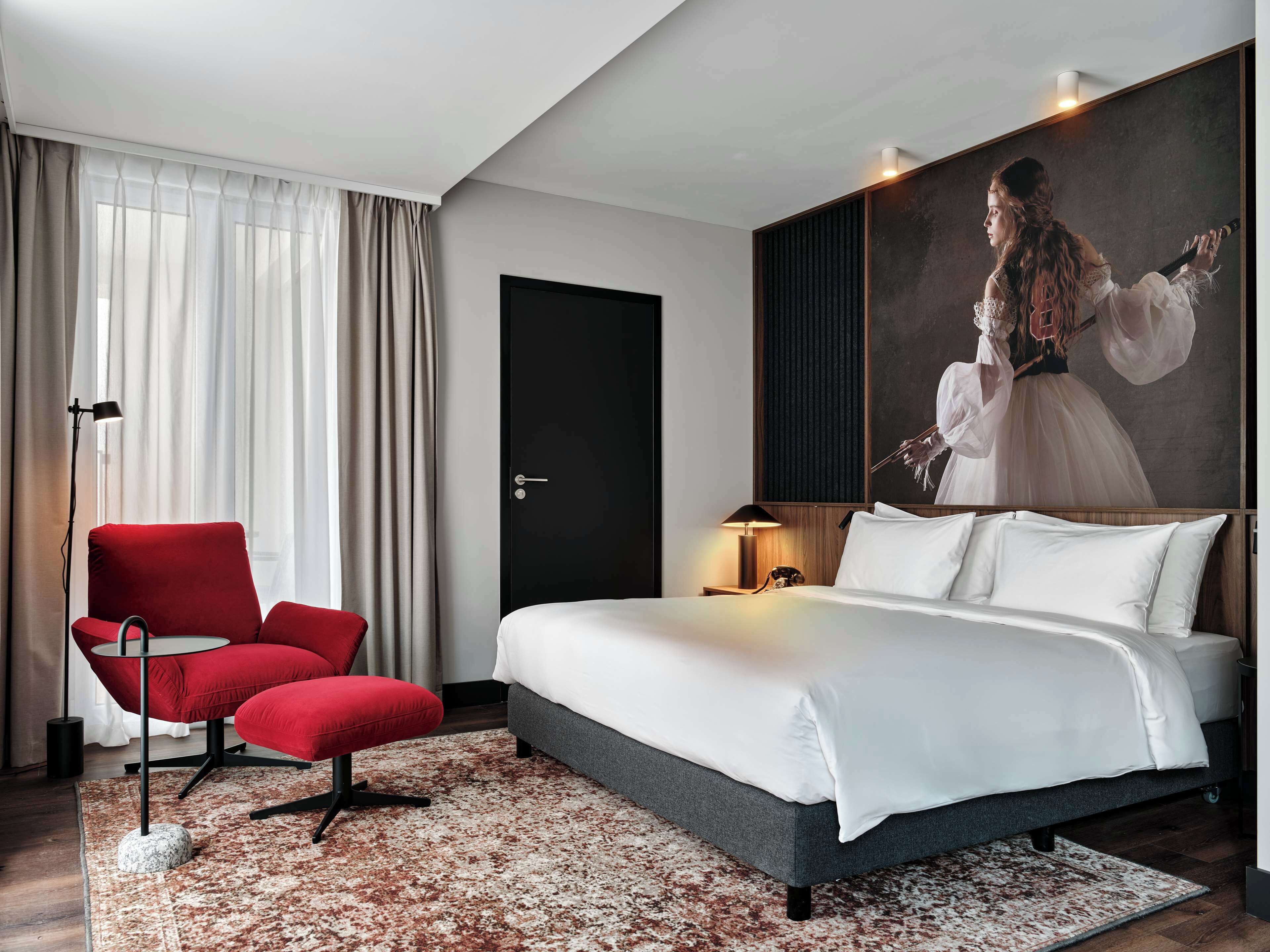 A modern hotel room with a large bed, a red armchair, a painting on the wall, and a floor lamp.