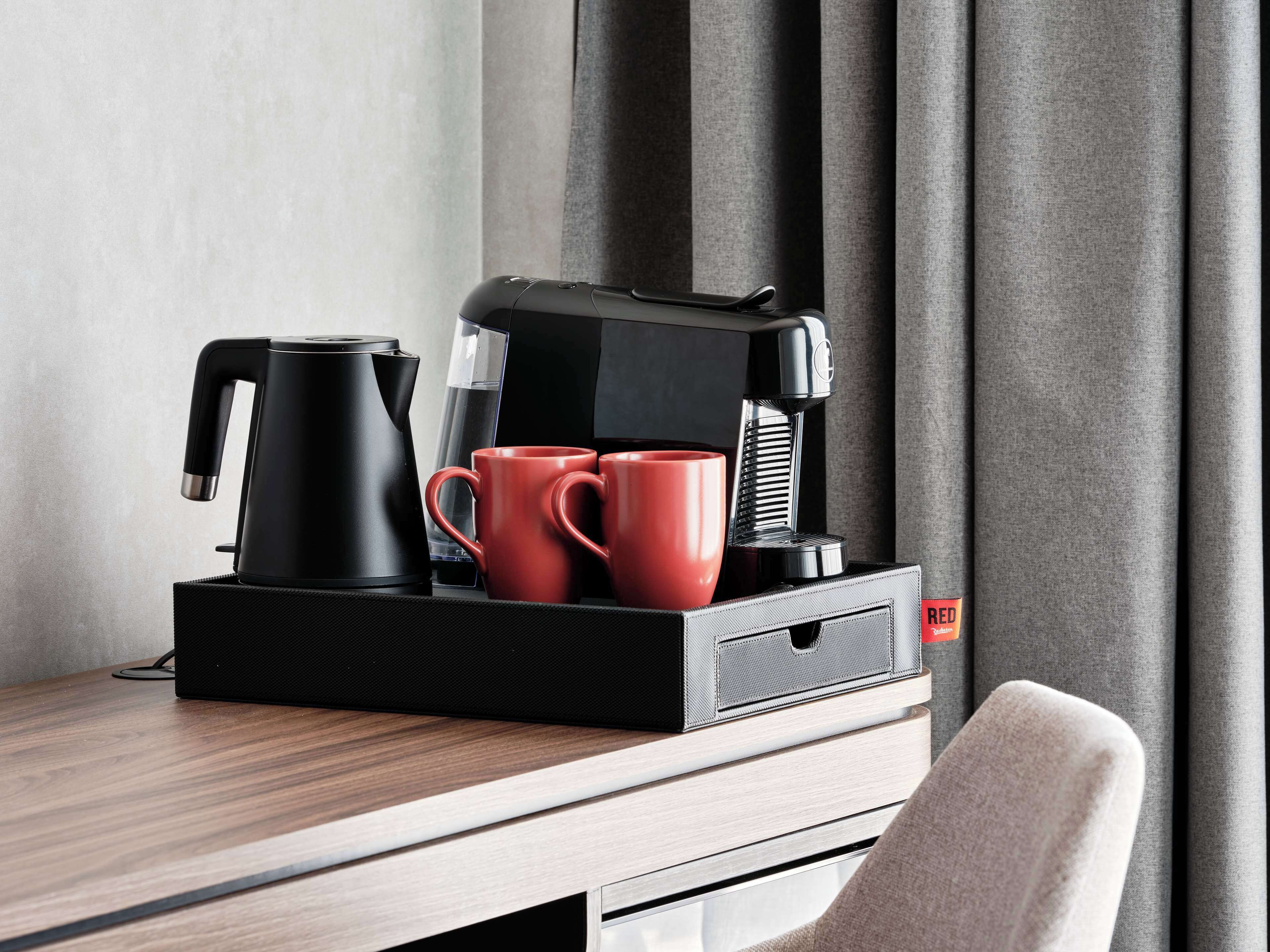 A modern coffee maker, two red mugs, and a clear glass on a wooden table in a hotel room.