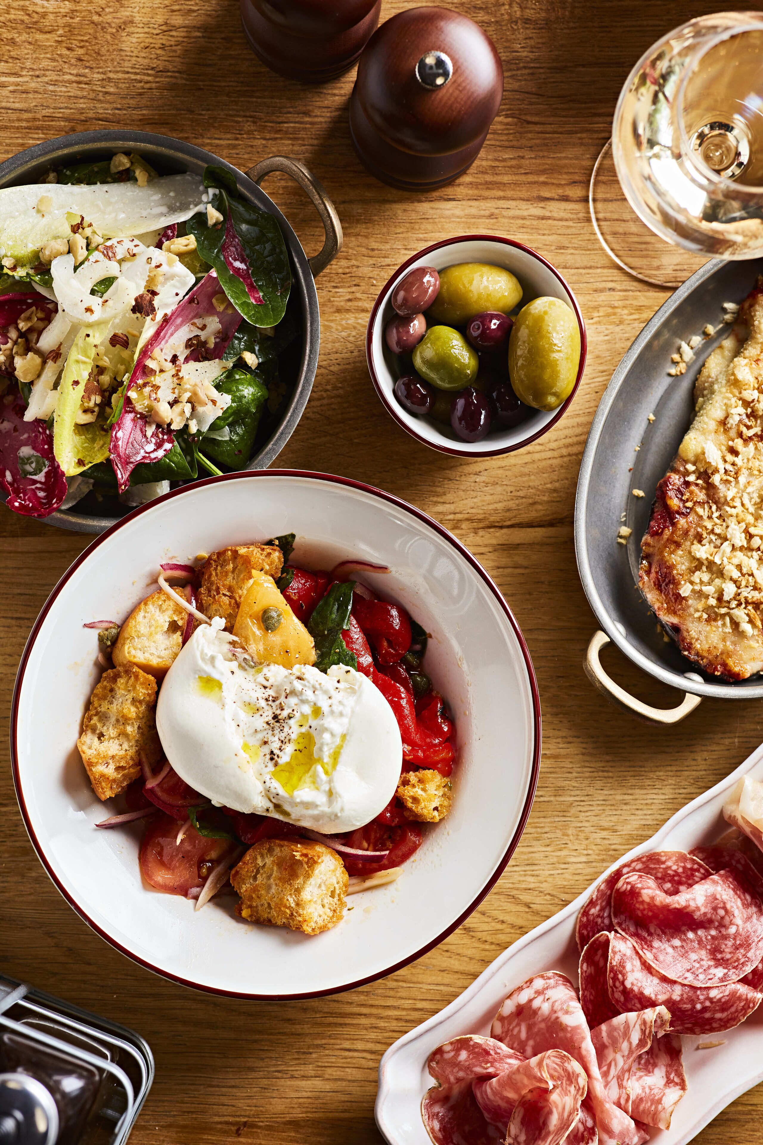A wooden table with various dishes including a salad, a poached egg, olives, and cured meats.