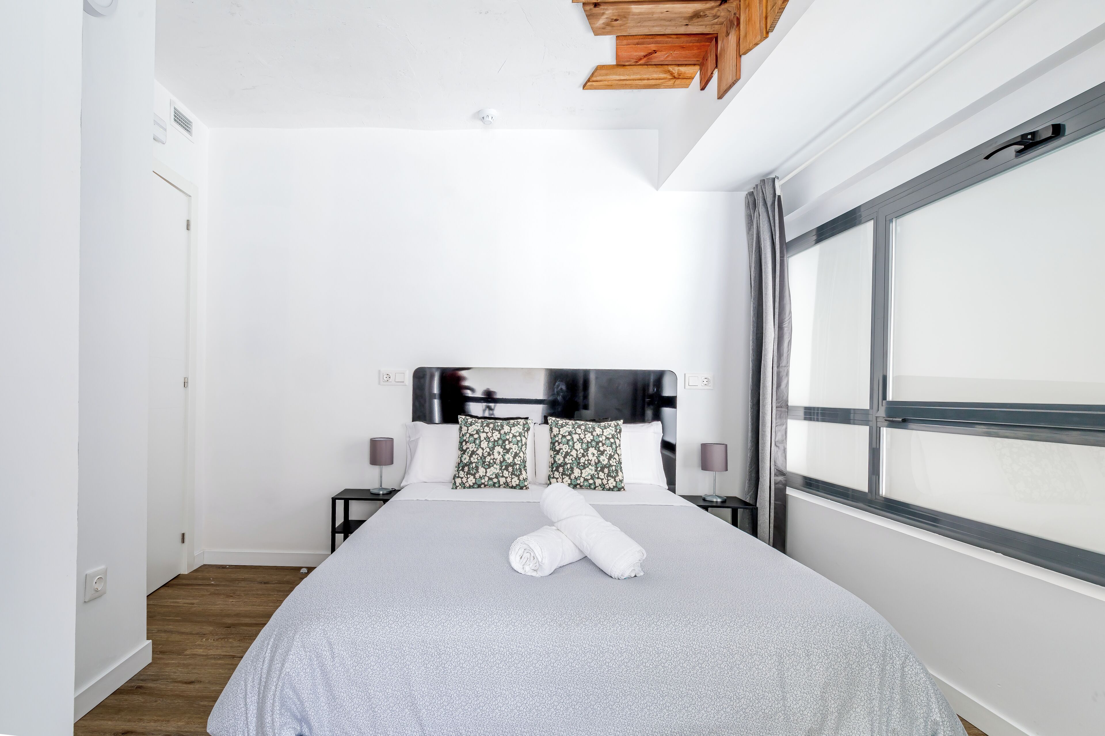 A neatly made bed with a grey bedspread, two floral pillows, and a rolled towel on the bed. There is a large window on the right side of the room.