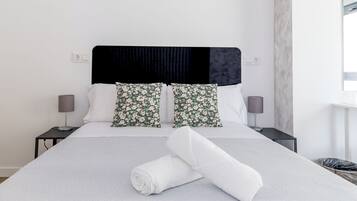 A neatly made bed with a white duvet, two floral pillows, and a black headboard. There are two bedside tables with lamps and a small trash bin.