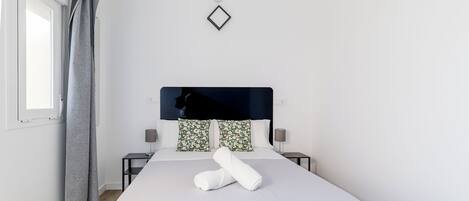 A modern hotel room with a large bed, two bedside tables, a nightstand, a window with curtains, and a wall-mounted light fixture.