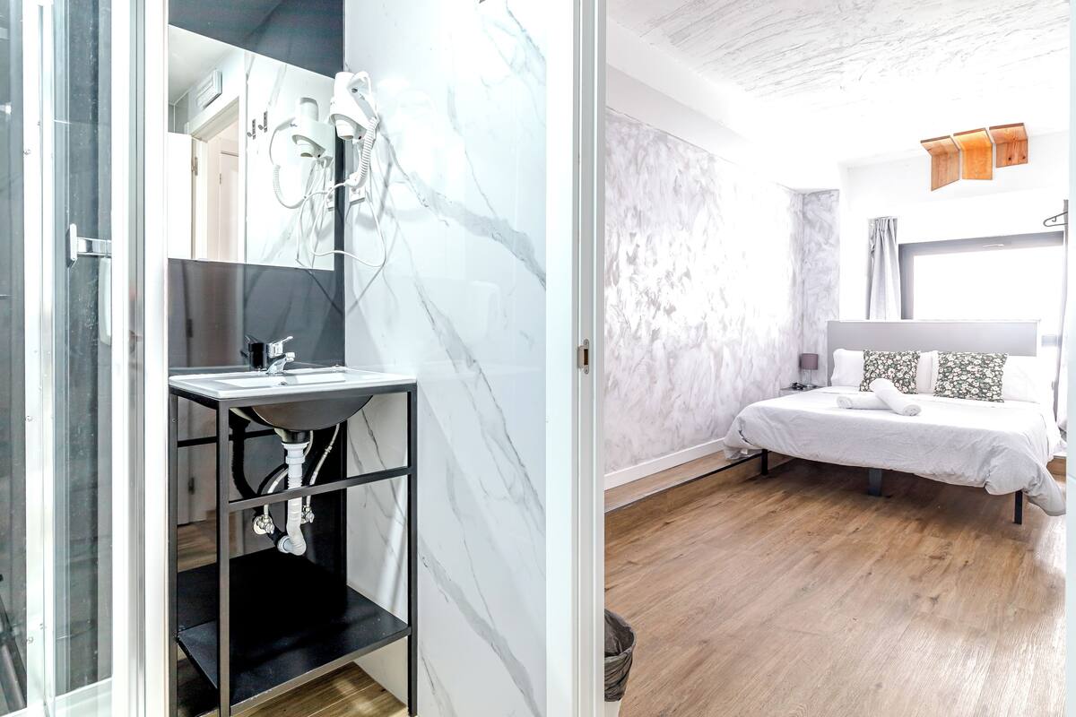 A bathroom with a marble sink, shower, and a glass-enclosed shower area. The bedroom features a bed with a white bedspread, a wooden floor, and a large window.