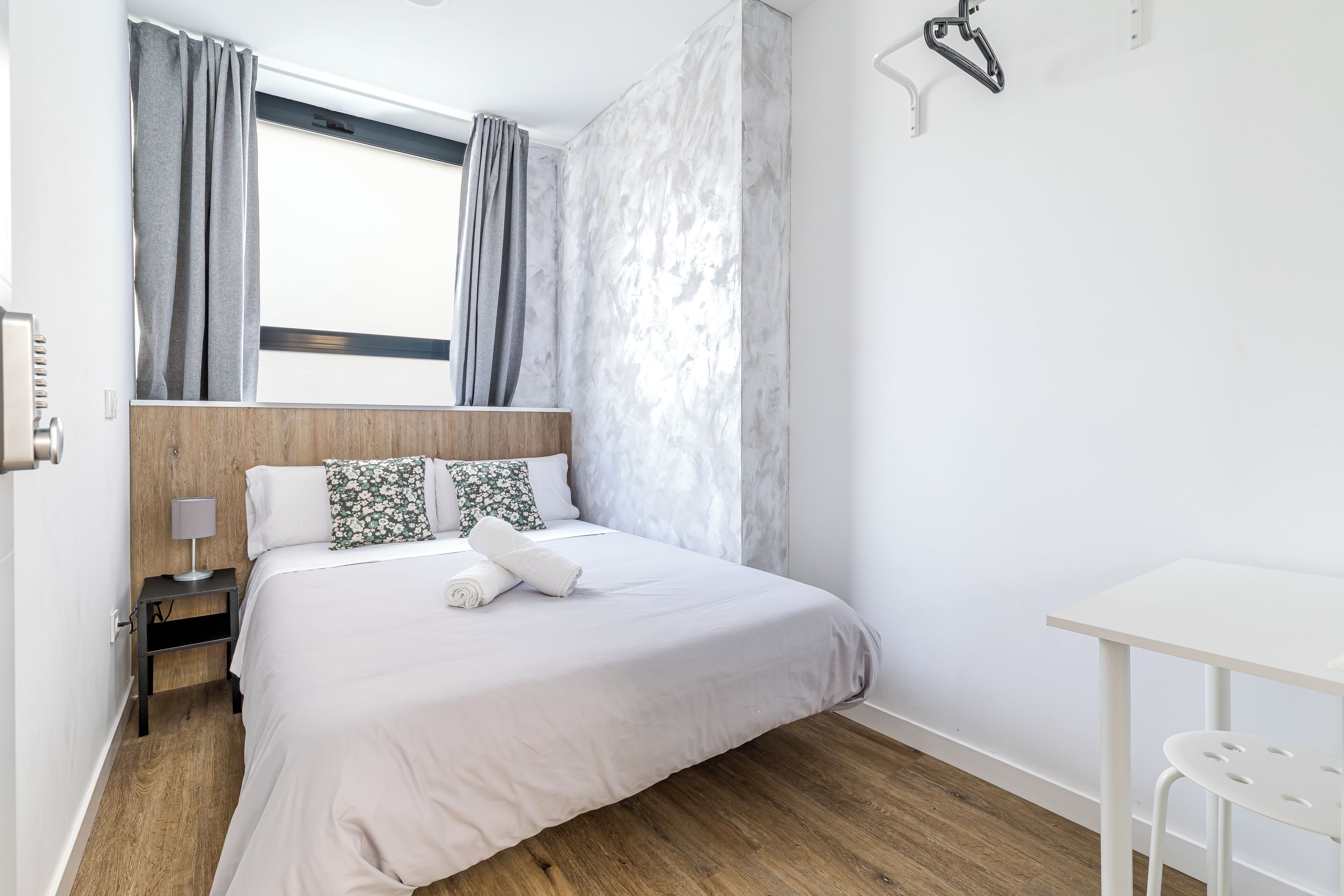 Un dormitorio moderno con cabecera de madera, cama con ropa de cama blanca, mesita de noche pequeña con lámpara y escritorio blanco con silla.