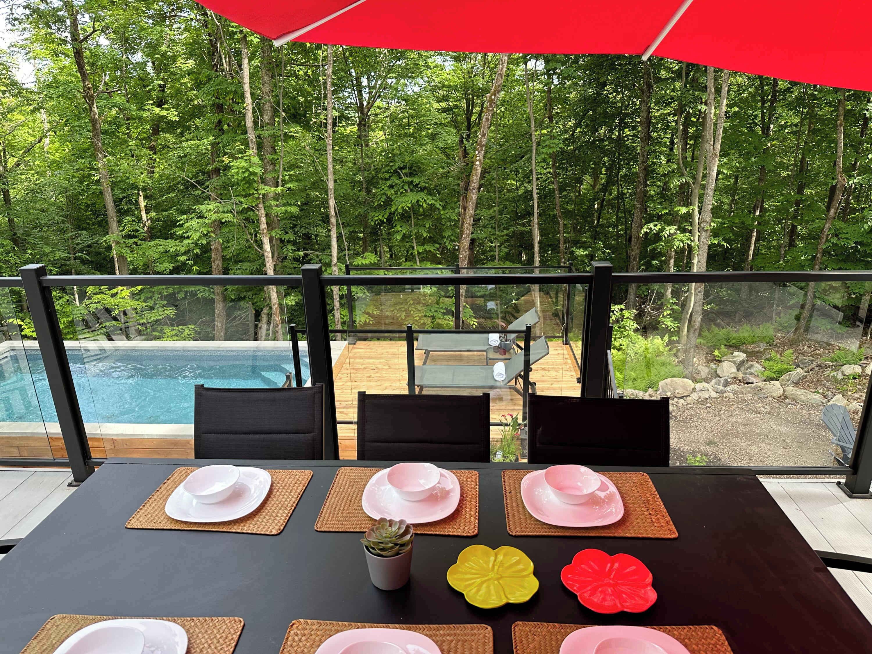 Une table à manger dressée avec des assiettes et une petite plante en pot, donnant sur une piscine et une zone boisée.