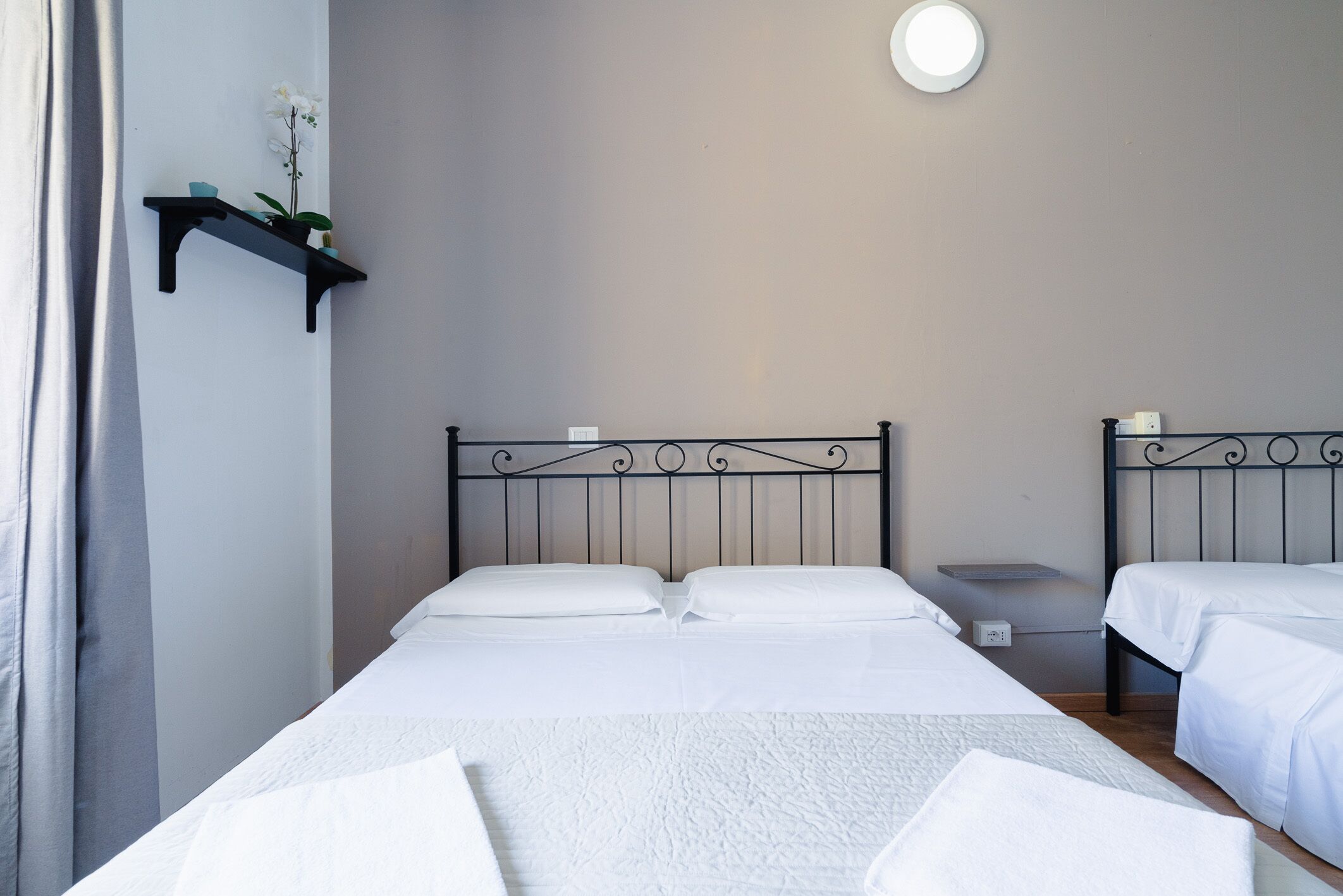 Une chambre avec deux lits, une table de chevet, une étagère murale ornée d’une plante et un luminaire au plafond.