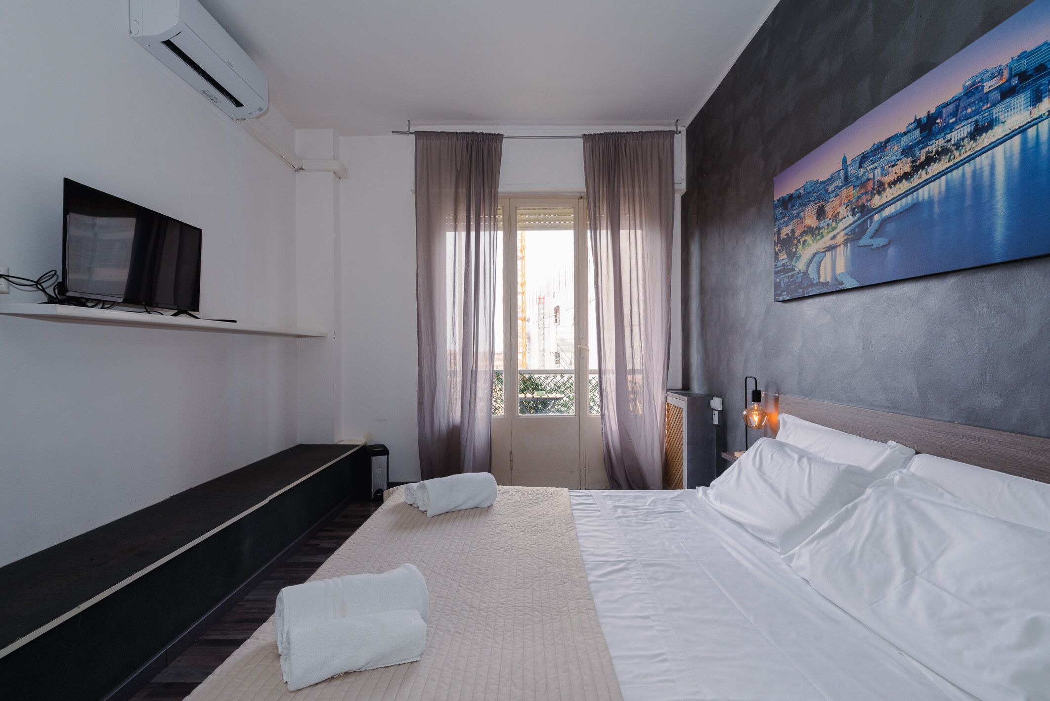 Una camera d'albergo con un letto, una TV su uno scaffale, un quadro al muro e una finestra con tende.