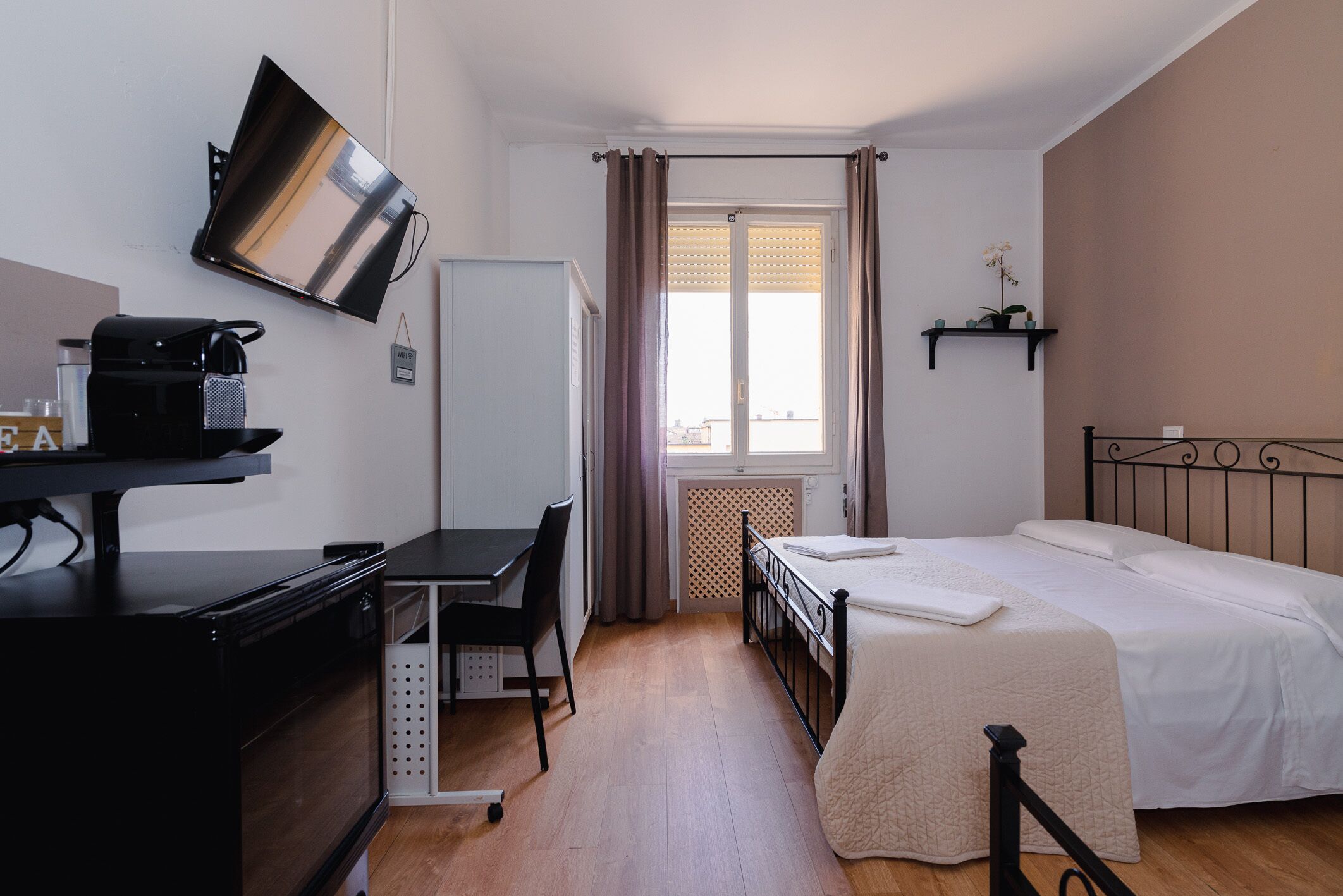 A compact hotel room with a bed, a desk with a computer, a refrigerator, and a TV mounted on the wall.