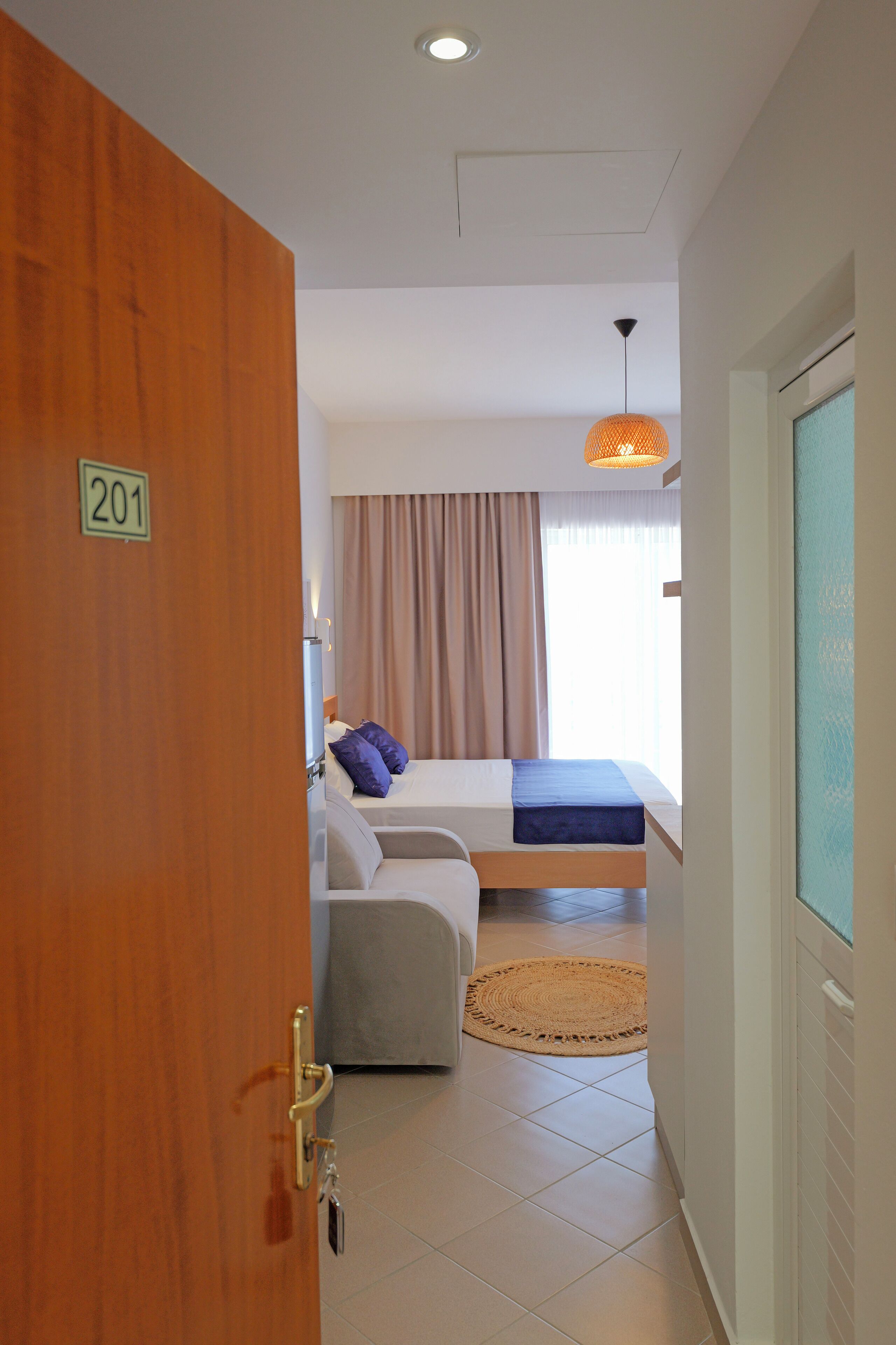 Habitación de hotel con cama, escritorio, silla, ventana con cortinas, lámpara en el techo y puerta con un número.