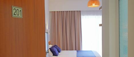 Habitación de hotel con cama, escritorio, silla, ventana con cortinas, lámpara en el techo y puerta con un número.