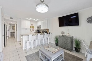 Une cuisine moderne avec des meubles blancs, un coin repas avec des chaises blanches, un canapé gris et un téléviseur à écran plat fixé au mur.