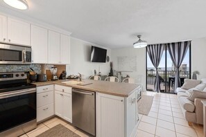 Una cocina moderna con armarios blancos, microondas, estufa y lavavajillas. Cuenta con una zona de comedor con mesa y sillas, y una sala de estar con sofá.