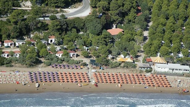 An aerial view of a beach with rows of beach umbrellas and sun loungers, a sandy beach, and a few buildings in the background. - Grotta dell' Acqua (Peschici)
