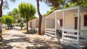 A row of single-story cabins with white exteriors and wooden shutters, surrounded by trees and a gravel path. - Grotta dell' Acqua (Peschici)