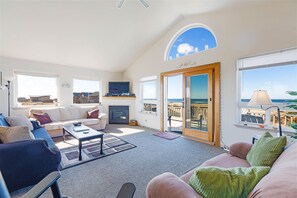 Relax in a spacious living area with a high ceiling, sofa, armchairs, and a fireplace, plus a sliding glass door to a balcony with sea views.