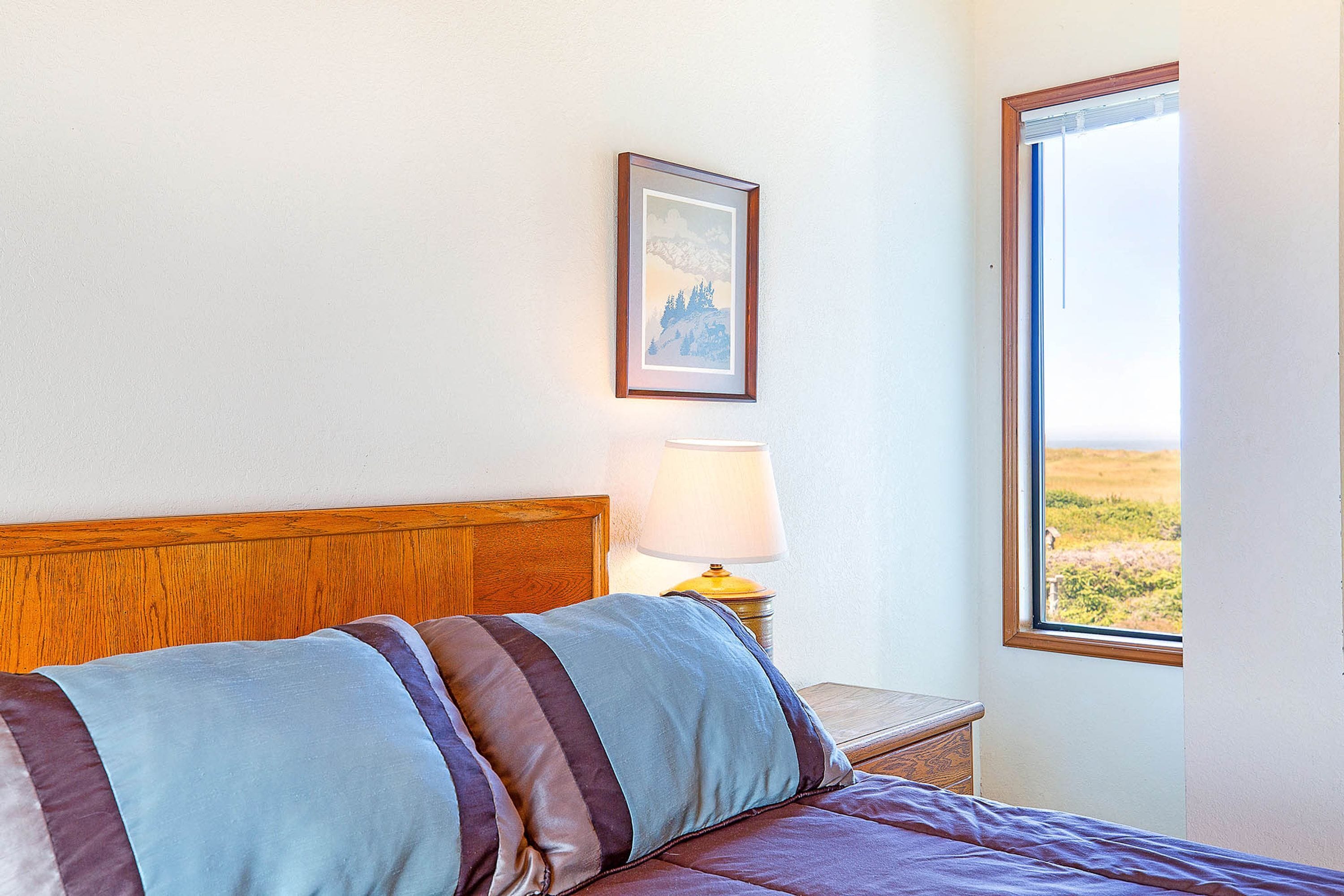A hotel room with a bed, a nightstand with a lamp, a framed picture on the wall, and a window with a view of the outdoors.