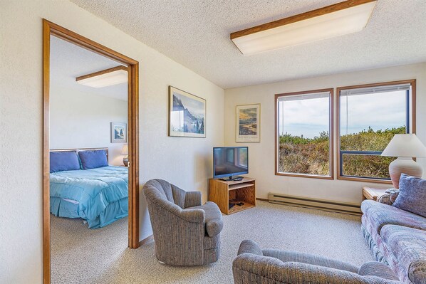 A room with a bed, a TV on a wooden stand, a sofa, a chair, and large windows with a view of greenery. - Haven House on Copalis Beach Near Ocean Shores (Copalis Beach)