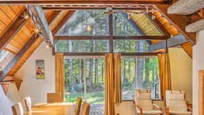 A dining area with a wooden table and chairs, large windows with curtains, and a view of a forest. - Four Stone Lodge Near Mt Rainier (Enumclaw)