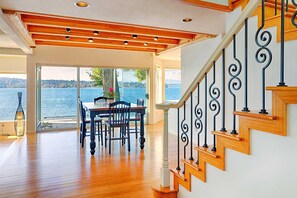 Una habitación amplia con una escalera de madera, una zona de comedor con mesa y sillas, y un gran ventanal con vistas a un cuerpo de agua.