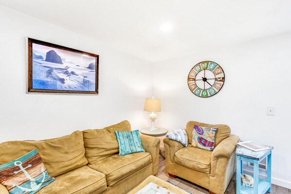 A living room with two beige sofas, a round wall clock, a framed picture, and a small side table with a lamp. - Shine On Condo in Ocean Shores (Ocean Shores)