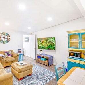 Un salon comprenant un canapé, un pouf, une table basse, une horloge, un téléviseur et un meuble contenant de la vaisselle.