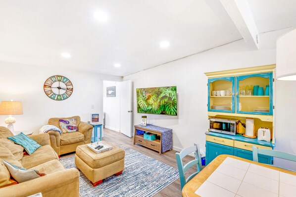 Un salon comprenant un canapé, un fauteuil, un pouf et une table basse. Un téléviseur est fixé au mur, il y a également une horloge et un meuble contenant des ustensiles de cuisine.