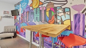 A room with a colorful mural, a wooden table, red chairs, and a bed with striped pillows.