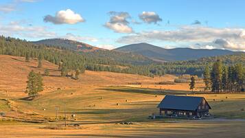 A rustic cabin with a large porch, surrounded by a grassy field with scattered hay bales and a backdrop of forested hills.